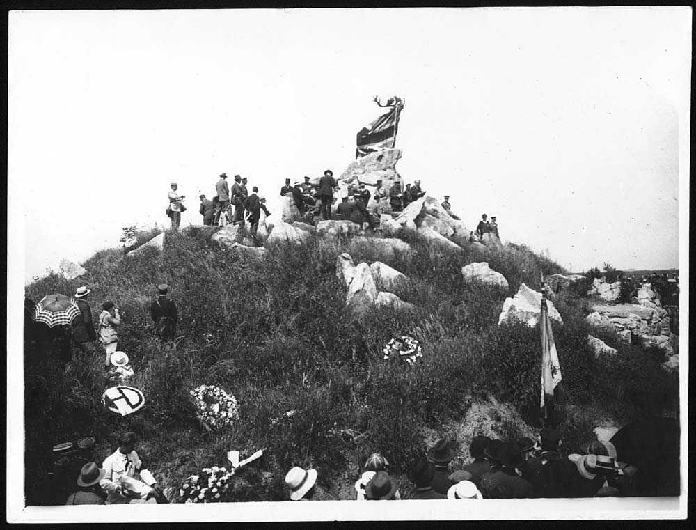 Photographie en noir et blanc – Des hommes retirent une bâche recouvrant une immense sculpture de caribou. Ils se trouvent sur une petite colline.